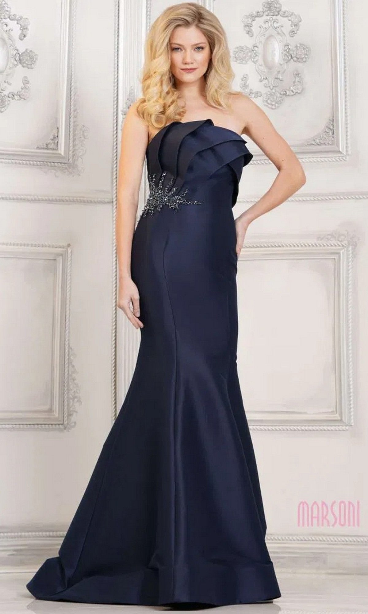 A woman stands before a decorative white wall in the navy Marsoni MV1308, a strapless mermaid long formal dress adorned with floral embellishments, showcasing its elegant design.